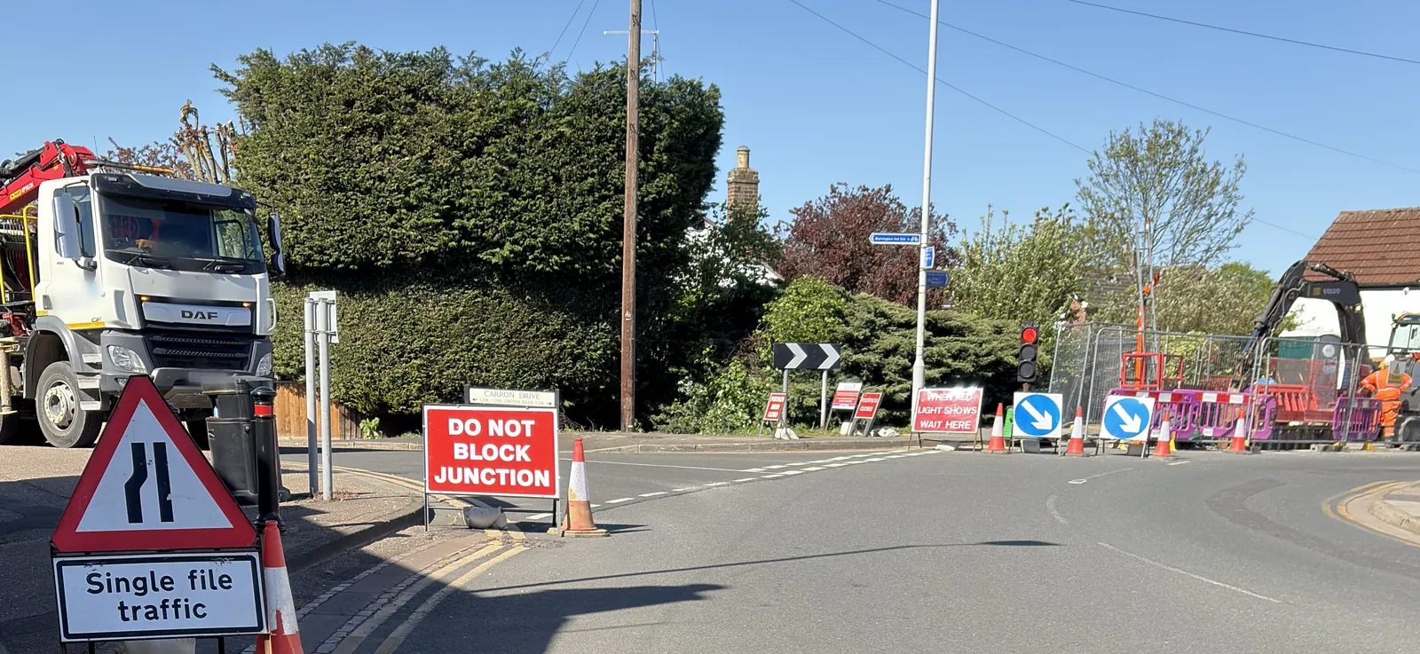 A road junction beside roadworks, with a lorry and a Single file traffic sign on the left, a red Do not block junction sign, a temporary traffic light showing red with a When red light shows wait here board, blue keep-left signs and a works compound beyond